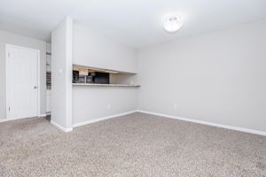 A spacious, empty room featuring light beige walls and plush carpet. On the left, a closed door leads to another area. To the right, an opening leads to a kitchen with a countertop and dark cabinetry visible. The room is well-lit by a ceiling light fixture.