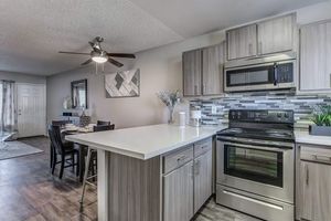 Modern kitchen with light wood cabinetry, stainless steel appliances, and a sleek countertop. A dining area is visible in the background with black chairs and a table. A ceiling fan is mounted above, and decorative elements like a vase and wall art add a stylish touch.