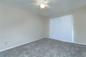 A spacious, empty room featuring light beige walls and plush gray carpeting. A ceiling fan is mounted in the center, and a large window with vertical blinds allows natural light to enter the space, creating a bright and airy atmosphere.