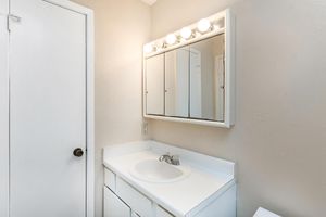 A simple bathroom featuring a white vanity with a circular sink, a large mirror with five overhead lights, and a closed door. The walls are a light neutral color, enhancing the bright and clean aesthetic.