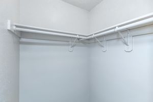 Empty corner closet with white walls and two horizontal white rods supported by brackets, creating space for hanging clothes or other items. The design is simple and minimalistic, emphasizing functionality in storage.