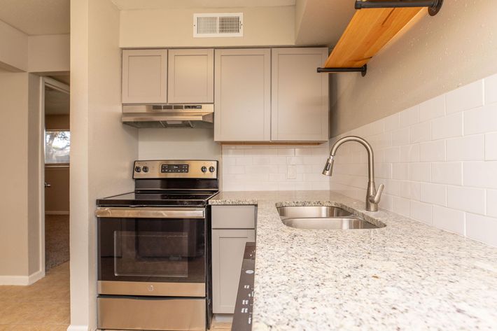 A modern kitchen featuring gray cabinetry, stainless steel appliances including an oven and hood, a double sink, and a spacious granite countertop. The backsplash is made of white subway tiles, and there is a wooden shelf above the counter for additional storage or decor. Natural light enters from an adjacent room.