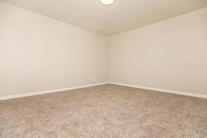 A vacant room with light beige walls and a carpeted floor. The space has a simple ceiling light fixture, and there are no furnishings or decorations, creating an empty and neutral atmosphere.