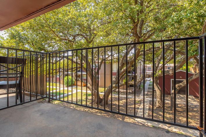 A view from a balcony showcasing a black metal railing, lush green trees, and a pathway leading to buildings in the background. The scene captures a peaceful outdoor area with sunlight filtering through the leaves, creating a serene atmosphere.