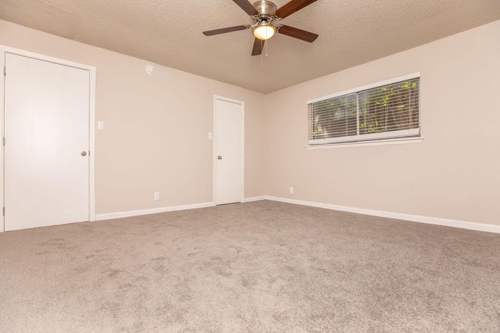 A spacious, empty room with light beige walls and a textured carpet. It features a ceiling fan, a closed door on the left, and a window with blinds allowing natural light to enter. The overall atmosphere is clean and inviting, perfect for personalization or decorating.