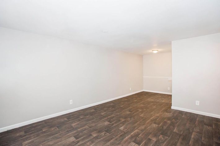 A spacious, empty room with light gray walls and dark laminate flooring. The ceiling is white with a central light fixture. There is a small transition area leading to another section of the room, with no furniture or decorations. The overall atmosphere is clean and modern.