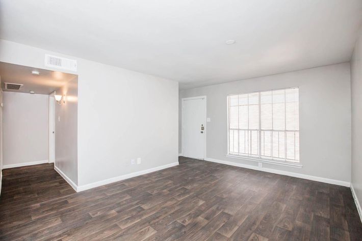 A spacious, empty room featuring light-colored walls, dark wood flooring, and a large window with blinds. There is a door on one side and a small entryway is visible. The overall atmosphere is bright and ready for furnishing.