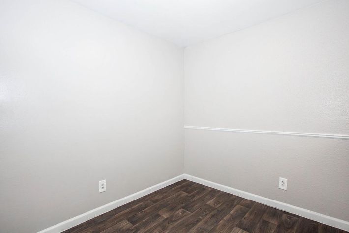 A sparsely furnished room with light gray walls and dark wood-like flooring. There is a white chair rail around the room. The space appears empty and ready for furnishing. There are two electrical outlets visible on the walls.