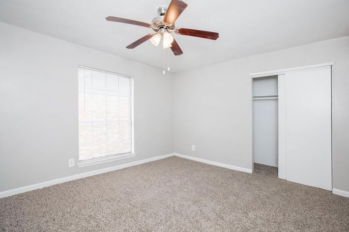 A clean, empty bedroom featuring light gray walls and soft carpet. A ceiling fan with three lights hangs from the ceiling. There is a window with white blinds allowing natural light, and a closet door on the right side. The room is spacious and well-lit, suitable for various decorating styles.