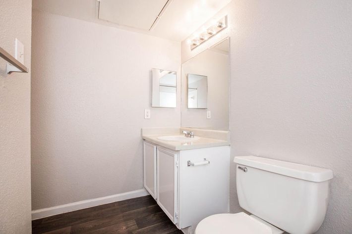 A clean and modern bathroom featuring a white toilet, a light-colored vanity with a sink, and two mirrors above the sink. The walls are painted in a light gray shade, and the floor has dark wood-like tiles. Bright overhead lights illuminate the space.
