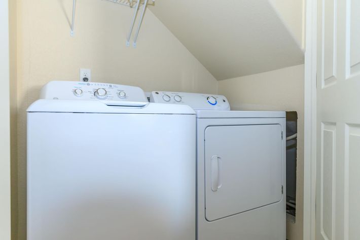 A washing machine and dryer stacked against a wall in a small laundry area. The appliances are white, with control panels visible on the front. The space is enclosed, with light-colored walls and a door partially visible.