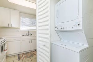A clean laundry area featuring a stacked washer and dryer, positioned next to a kitchen with white cabinetry. The space includes a sink with a window above it, and a colorful patterned rug on the tiled floor. Light brightens the room through the window, creating an inviting atmosphere.