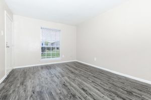 A vacant interior room with light beige walls and a medium-colored wood-like floor. A window with blinds allows natural light in, showcasing a view of greenery outside. The space is unadorned, creating a minimalist and open atmosphere.