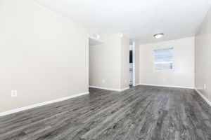 A spacious, empty living area featuring light beige walls and dark wooden laminate flooring. A single ceiling light fixture illuminates the room, which has a window providing natural light. There is a doorway leading to another space, creating an open and inviting atmosphere.