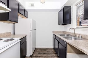 A modern kitchen featuring black cabinetry, a white refrigerator, a white stove, and a sink with a chrome faucet. The countertops are light-colored and the flooring is dark. Natural light enters through a window, creating a bright and spacious feel.