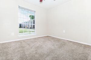 A minimally furnished room with light beige walls and a large window featuring white blinds. The floor is covered in soft beige carpet, and the space is empty, creating a bright and inviting atmosphere.