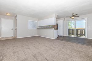 Spacious living area with beige carpet, adjacent kitchen featuring a breakfast bar, light-colored walls, and a ceiling fan. Large windows with blinds allow natural light, and sliding glass doors open to a balcony. The front door is visible on the left, adding to the welcoming atmosphere.