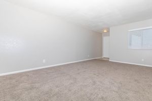 A spacious, empty room with light gray walls and a pale carpet. A single window with white blinds lets in natural light. The ceiling is simple, and there is a light fixture visible. The entrance to another area is seen in the background, creating a clean and minimalist aesthetic.