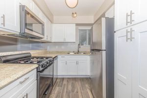 A modern kitchen featuring white cabinetry, a stainless steel refrigerator, and a microwave above the gas stove. The countertop is light-colored granite, and there are neutral wall colors. Natural light enters through a window above the sink, with wood-like flooring completing the contemporary look.