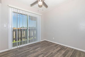 A bright, empty room with beige walls and a wood-like floor. There are vertical blinds covering a sliding glass door leading to a balcony, with a ceiling fan above. The space is well-lit, giving a spacious and airy feel.