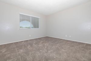A spacious, empty room with gray carpet and light beige walls. A window with white blinds allows natural light to fill the space, framed by a curved wall. The floor is clear of furniture, emphasizing the room's openness and potential for personalization.