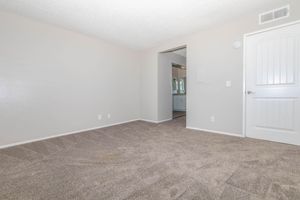 A spacious, empty room with light gray walls and beige carpet. There is an open doorway leading to another room, with a mirror and additional lighting visible in the background. The overall atmosphere is bright and airy, suitable for various interior design possibilities.