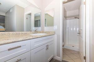 A clean bathroom featuring a granite countertop with dual sinks, white cabinetry, a mirror, and a small shower area. The walls are painted in neutral tones, and there is a tile floor. The overall atmosphere appears bright and well-maintained, with a glimpse of a larger living space visible through an open door.