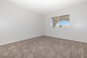 A vacant room with light gray walls and carpet. There is a window with white blinds allowing natural light to enter. The floor is clean and the space is uncluttered, creating a simple and open atmosphere.