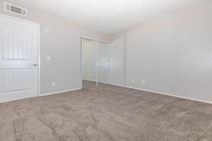 A neat, empty room featuring beige walls and tan carpet. A door leads to the room, and a large mirrored closet on the opposite wall reflects the room's light. The space is well-lit, with no furniture present, giving it a clean and open appearance.