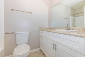 A clean and modern bathroom featuring a white toilet, a white vanity with a granite countertop, and a large mirror. The space is well-lit, with tiled flooring and a towel rack on the wall. A glass shower enclosure can be seen in the background, providing a sleek and functional design.
