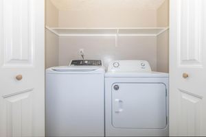 A laundry room with a washing machine on the left and a dryer on the right, both white. The appliances are set against a light gray wall with a shelf above them and white doors on either side, creating a neat and organized appearance.