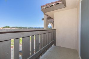 A view from a small balcony or patio with a wooden railing, featuring a clear blue sky and mountainous landscape in the background. The architectural style is modern, with a flat roof and stucco walls. The scene suggests a peaceful, outdoor space for relaxation.