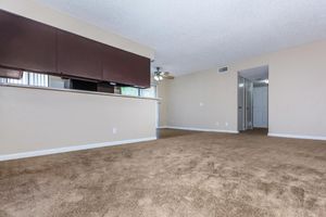 An indoor living space featuring beige walls and a light brown carpet. There is a partial view of a kitchen with dark cabinetry in the background. A ceiling fan is visible, and a door leads to another room. Natural light comes through a window, creating a bright and open atmosphere.