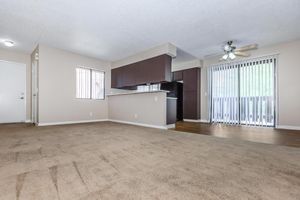 A spacious, unfurnished living area featuring light beige carpet, neutral walls, and large windows with vertical blinds. A kitchen area is visible with dark cabinetry, and a ceiling fan is installed. The layout suggests a comfortable, open-concept design, ideal for various furniture arrangements.