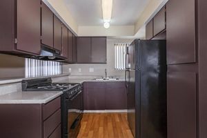 A modern kitchen featuring dark wooden cabinets, a black gas stove, and a black refrigerator. The countertop is light-colored, and there is a sink under a window with blinds. The wooden floor adds warmth to the space, and the room is well-lit with overhead lighting.