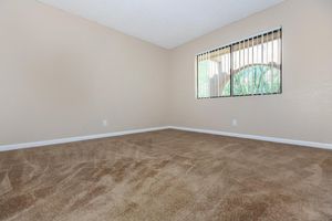 Empty room with beige walls and carpeted flooring. A window with vertical blinds allows natural light to enter, creating a bright and open atmosphere. The space is clean and uncluttered, suitable for various uses or decoration.