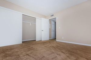 A simple, empty room featuring beige walls and a light brown carpet. There are two doors: one leads to a small closet with a white sliding door, and the other opens into a hallway. The room has no furniture, creating a minimalistic and spacious feel. Natural light may be entering from an unseen source.