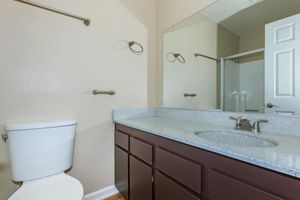 A clean bathroom featuring a white toilet, a double sink vanity with a grey countertop, and a large mirror above. The walls are a soft beige color, and there is a shower area visible in the background, along with towel bars mounted on the wall. The overall design is modern and minimalistic.