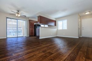 Spacious, unfurnished living area featuring a wooden floor, ceiling fan, and natural light from large windows. The open floor plan includes a kitchen with dark wood cabinetry partially visible. Sliding glass doors lead to an outdoor space, while the entrance door is located on the right side of the room.