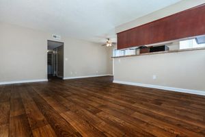 A spacious, light-filled living room featuring wooden floors, a ceiling fan, and warm beige walls. The open layout connects to a kitchen area with a low partition. A doorway leads to another room, enhancing the sense of space and comfort.