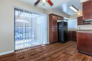 Bright kitchen interior featuring wooden flooring, dark cabinetry, and a black refrigerator. A window with vertical blinds allows natural light, revealing a view of a car outside. The kitchen includes a countertop and overhead lighting, creating a warm and inviting atmosphere.