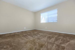 Empty room with beige walls and a light beige carpet featuring a subtle pattern. A window with white blinds allows natural light to enter, illuminating the space. The room has no furniture, emphasizing its spaciousness and neutral color scheme.