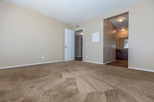 A spacious, empty room with beige walls and light brown carpet. A white door leads to another room, while a doorway on the right opens into a bathroom area. The room is well-lit, featuring ceiling lights. The overall atmosphere is clean and neutral, suitable for various decorating styles.