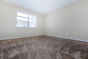 Empty room with beige walls and carpeted floor. A window with white blinds allows natural light to enter. There are no furnishings or decorations present, emphasizing the spaciousness of the room. The light creates subtle shadows on the carpet, enhancing the overall simplicity and neutrality of the space.