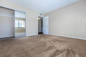 A spacious and empty room with beige walls and a light brown carpet. There is a large mirror closet on one side, and a doorway leading to another room can be seen in the background. Natural light enters through the window, creating a bright and inviting atmosphere.