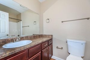 A clean bathroom featuring a countertop with a sink, a large mirror, a toilet, and a towel rack. The walls are painted a light color, and there is a door leading to another area. The flooring appears to be hardwood.