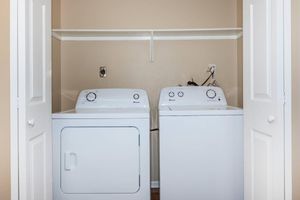 A laundry room featuring a white washing machine and a white dryer side by side. The machines are located in a small space with beige walls and a shelf above. The doors of the room are open, revealing the appliances organized neatly.