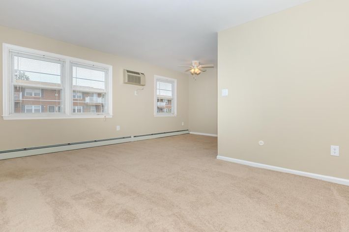 A spacious, empty room with light beige walls and a tan carpet. There are three windows allowing natural light, and a ceiling fan is visible. An air conditioning unit is installed on one wall, while another wall features an electrical outlet. The room has a clean, simple aesthetic.