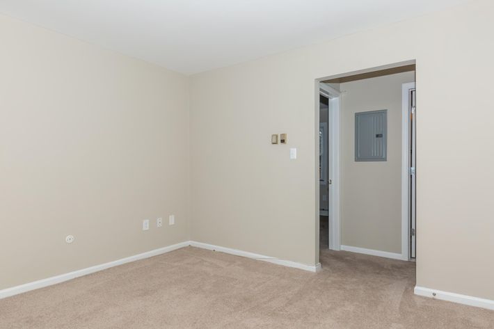 Empty room with beige walls and light brown carpet. A doorway on the right leads to another space, while the left wall features two light switches. There are no furnishings or decorations in the room, creating a spacious and minimalist appearance. Natural light may enter from the adjacent area.