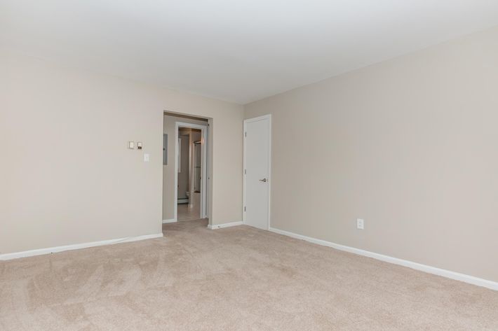 Empty room with beige walls and tan carpet. There is a doorway on the left leading to another area, and a closed door on the right. The room is well-lit with soft natural light, giving it a spacious and clean appearance. Ideal for use as a living space or office.